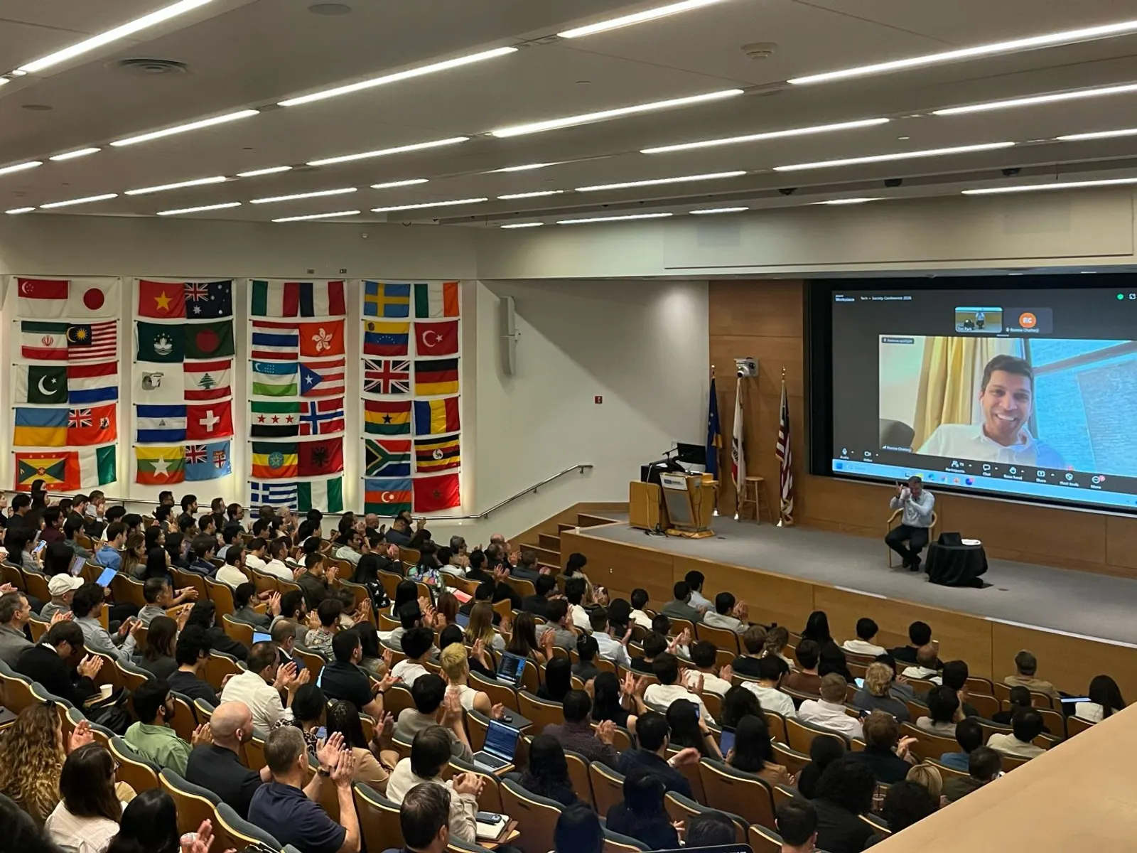 A packed lecture hall applauding a speaker who joins by video call on the big screen, with a moderator seated on stage beside it